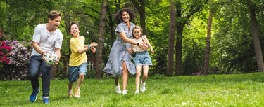 En familie leger sammen i en grøn park. Forældre og børn løber og smiler, mens de spiller fodbold. Børn bruger Xplora smartwatches, som symboliserer aktiv leg og sikker forbindelse.