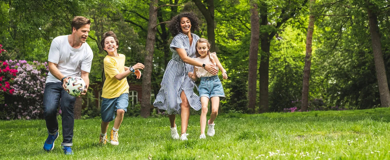En familie leger sammen i en grøn park. Forældre og børn løber og smiler, mens de spiller fodbold. Børn bruger Xplora smartwatches, som symboliserer aktiv leg og sikker forbindelse.