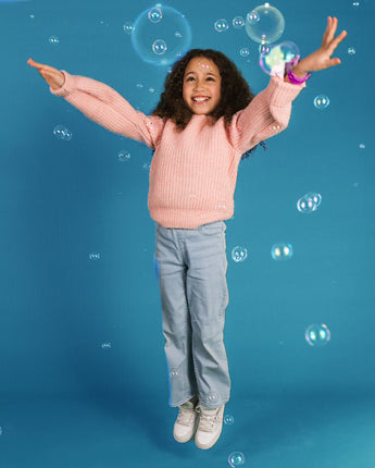 En glad pige med krøllet hår hopper i luften med armene udstrakt, omgivet af sæbebobler. Hun har en lyserød strikket sweater, lyserøde jeans og hvide sneakers på. På håndleddet har hun et lyserødt smartwatch.