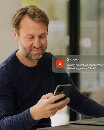 Forælder kigger på sin mobil derhjemme med en Xplora-notifikation om, at barnet er ankommet til en tryg zone.